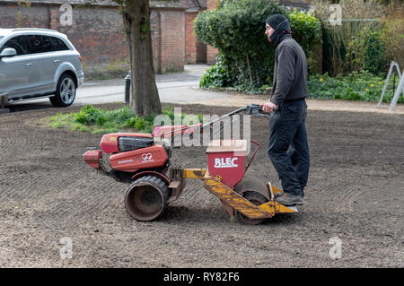 Winchester, Hampshire, Inghilterra England Regno Unito. Marzo 2019. Paesaggio giardiniere a cavallo su un distributore di seme e il rullo per formare un nuovo prato. Foto Stock