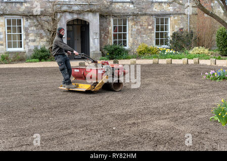 Winchester, Hampshire, Inghilterra England Regno Unito. Marzo 2019. Paesaggio giardiniere a cavallo su un distributore di seme e il rullo per formare un nuovo prato. Foto Stock