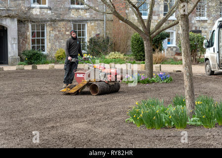 Winchester, Hampshire, Inghilterra England Regno Unito. Marzo 2019. Paesaggio giardiniere a cavallo su un distributore di seme e il rullo per formare un nuovo prato. Foto Stock