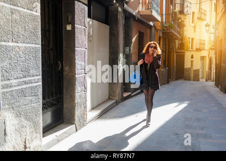 Donna che indossa gli occhiali da sole a parlare su smart phone mentre si cammina in vicolo nel mezzo di edifici in città Foto Stock