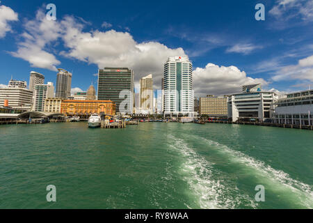 La vista dal retro di un traghetto come la barca lascia il porto di Auckland, con lo skyline della città sullo sfondo contro un luminoso cielo blu. Foto Stock