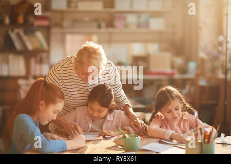 Ritratto di donna matura lavora con un gruppo di bambine pittura in tecnica di classe, scena illuminata da luce solare sereno, spazio di copia Foto Stock