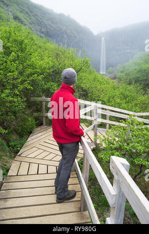 L'uomo traveler in giacca rossa in piedi su un ponte di legno. Nuvoloso mattina d'estate. Skaftafell National Park, Svartifoss cascata, Islanda, l'Europa. Natura Foto Stock