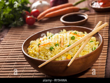 Cuocere il riso bianco con curry e verdure in una ciotola con le salse e i bastoncini su una tavola di legno di bambù marrone dello sfondo. Foto Stock