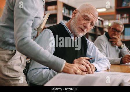 Vista laterale di un docente aiutare un anziano uomo in aula mentre un altro studente guarda a. Docente di spiegare i concetti dal libro di un anziano Foto Stock