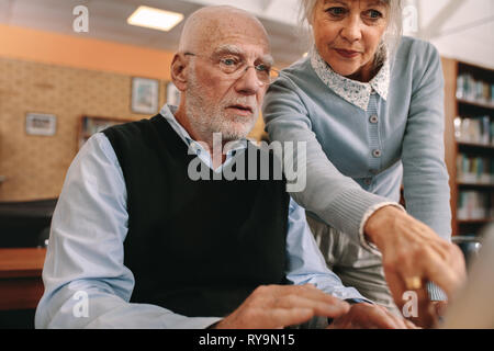 Senior donna rivolta verso lo schermo di un computer mentre un uomo anziano guarda. Coppia senior presso una libreria utilizzando un computer. Foto Stock