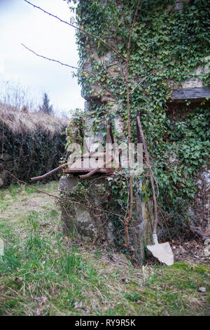 Vecchie e arrugginite carriola e la pala su una parete di una casa ricoperta con poison ivy nel villaggio abbandonato Slapnik nella regione Goriška Brda, Slovenia. Foto Stock