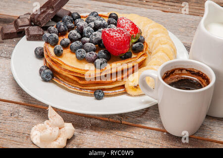 Una sana prima colazione, caffè e deliziosi pancake con frutti di bosco freschi e banana, su un tavolo di legno. Foto Stock