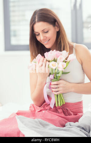 Bella donna attraente seduta sul letto teneramente tenendo in mano un mazzo di fiori di primavera tulipani. Foto Stock