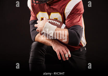 Giocatore di football americano seduto in una posizione di predisposizione, le mani per tenere una mache su uno sfondo nero, concept Foto Stock