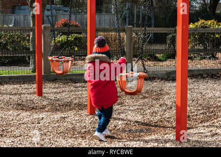 Due anni di vecchio ragazzo giocando con altalene nel parco indossando un bobble hat e rivestimento rosso Foto Stock