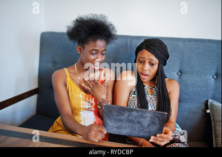 Due africano nero fidanzate in abiti estivi in seduta cafe, guardando lo spazio vuoto piastra con facce scioccante. Foto Stock