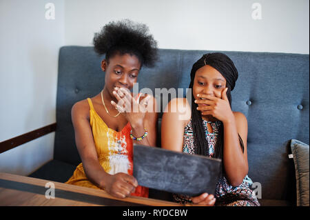 Due africano nero fidanzate in abiti estivi in seduta cafe, guardando lo spazio vuoto piastra con facce scioccante. Foto Stock