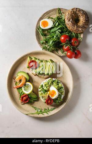 Varietà di panini vegetariani con fette di avocado, pomodori secchi, uovo, gamberetti, rucola servita su piastra in ceramica oltre bianco grigio maculato backgr Foto Stock