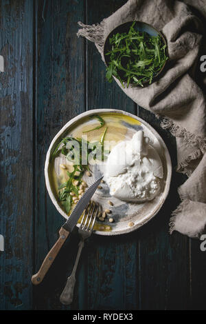 Italiano a fette di formaggio burrata, fresca insalata di rucola, pinoli e olio d'oliva in ceramica bianca piastra sul panno scuro su tavola in legno sfondo. La piatta Foto Stock