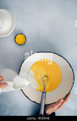 Donna versando il latte in uova sbattute in una grande ceramica recipiente di miscelazione. Foto Stock