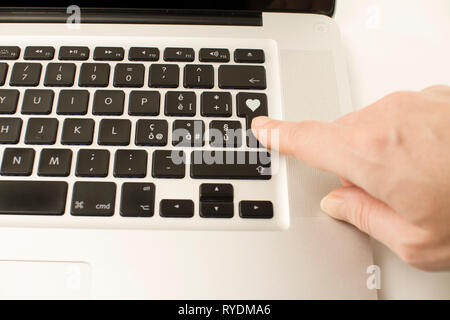 Anche la tecnologia si applica ai rapporti affettivi: una donna di mano è circa alla pressione di un tasto sulla tastiera del notebook con un cuore impresso Foto Stock