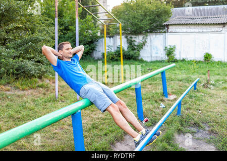 Montare l'uomo facendo allenamento calisthenics sgranocchia sit ups sulle barre parallele orizzontali esercizio nel parco all'aperto in Rivne, Ucraina parco giochi Foto Stock