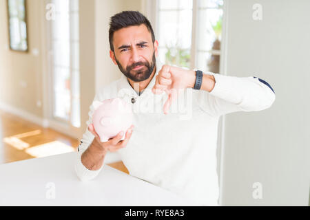 Bel uomo ispanico holding salvadanaio con volto arrabbiato, segno negativo che mostra antipatia con il pollice verso il basso, il concetto di rifiuto Foto Stock