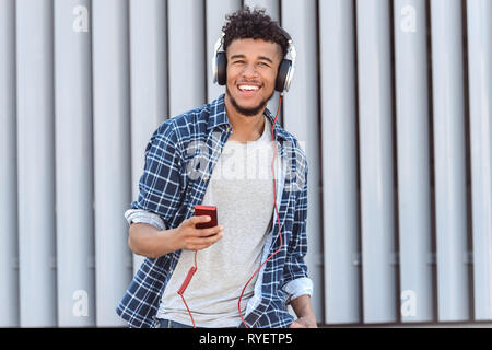 Per il tempo libero all'aria aperta. Mulatto guy nelle cuffie in piedi isolato su una strada di città ascoltando musica sul lettore toothy sorridente Foto Stock