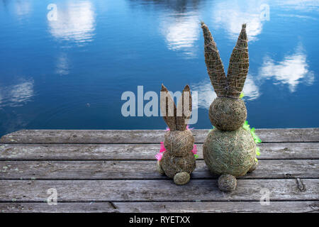 La paglia figura Bunnys dal retro sul molo di pista-doga con sky riflessione sull'acqua a un lago di Pasqua e di primavera il concetto di sfondo Foto Stock