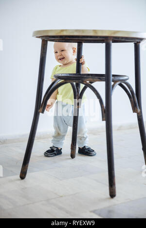 Carino bambina in piedi mentre la gamba di presa di uno sgabello e ridere. Bambino facendo i primi passi. Foto Stock