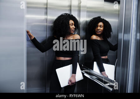 Moda giovane bella African American Business donna con acconciatura afro usura in elegante nero, stand ad ascensore con il portatile a mani. Foto Stock