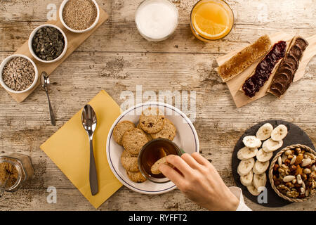 Una donna di immersione a mano il biscotto nel tè a un tavolo di legno fissato per una dolce colazione vegana shot dal di sopra con fette di banana, frutta secca mista, veg Foto Stock