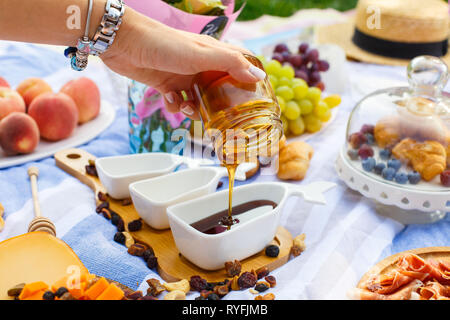 Donna mano versa il miele dalla bottiglia trasparente a sauceboat, a picnic estivo dello sfondo. Foto Stock