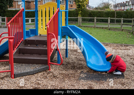 Due anni di vecchio ragazzo giocando sulla slitta in plastica presso il parco indossando un cappotto rosso e bobble hat Foto Stock