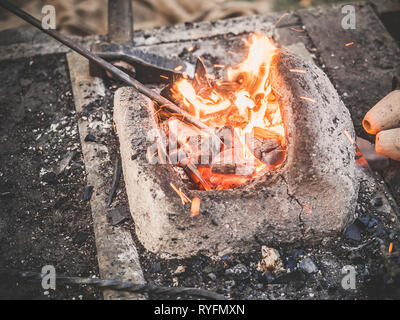 Il fabbro detiene la billetta sui carboni ardenti in un forno di argilla. fabbro riscaldamento ferro spada di metallo Fabbricazione di marching forge Foto Stock