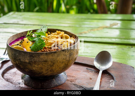 Colazione frullato ciotola sul tavolo in cafe con frutti assortiti, cereali e menta pronto a mangiare servita in un ambiente rustico cercando il cocco ciotola con un cucchiaio. Foto Stock