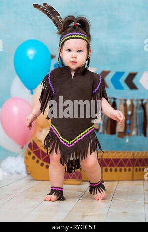 Poco carino ragazza in costume di Apache capo indiano e in un copricapo di piume si siede in canoa. I bambini di vacanza primo compleanno e smash torta 1 anno Foto Stock