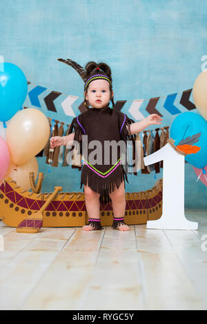 Poco carino ragazza in costume di Apache capo indiano e in un copricapo di piume si siede in canoa. I bambini di vacanza primo compleanno e smash torta 1 anno Foto Stock