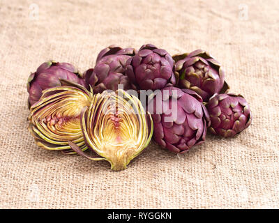 Carciofi crudi, Mediterraneo, vegetali non cotti su rustiche hessian. Un taglio aperta per vedere all'interno. Foto Stock