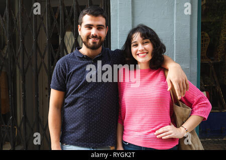 Felice e attraente coppia giovane in posa e sorridente alla fotocamera in strada contro uno sfondo di negozi Foto Stock