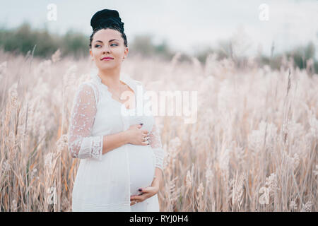 Incinta bella donna in pizzo bianco boho abito in piedi in canne e tenendo il suo ventre Foto Stock