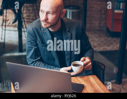 Il pensiero allettante successo per adulti calvo uomo barbuto in tuta con computer portatile in cafe Foto Stock
