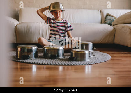 Futuro drummer boy indossando una ciotola sulla testa seduto sul pavimento e giocare su pentole come un batterista. Ragazzo di riproduzione di musica con pentole di cottura a0 Foto Stock