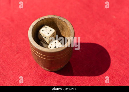 Vintage dadi di legno in una coppa di legno o pentola che sorge su di un soleggiato tavolo ricoperto liscio con panno rosso Foto Stock