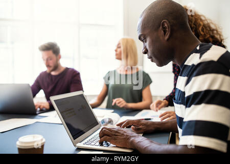 Giovani designer africani guardando i grafici sul suo computer portatile mentre si lavora con i colleghi attorno a un tavolo per riunioni in un ufficio Foto Stock