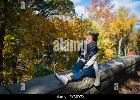 Giovane donna sorridente appoggiato su sfondo giallo di alberi e foglie. Felice elegante ragazza si siede da solo nel parco. Soleggiata giornata autunnale nella città di New York, Stati Uniti d'America. Foto Stock