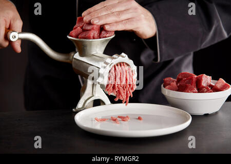 Cuocere in nero uniforme di macinazione delle carni bovine fresche o di maiale usando il vecchio manuale di metallo tritacarne sul banco di cucina. Pezzi di carni le carni rosse in bianco ciotola. Cooki Foto Stock