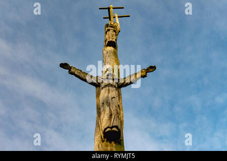 Rustico intagliati a mano la figura di Cristo con bracci estesi su un albero guardando dal basso contro un nuvoloso cielo blu Foto Stock