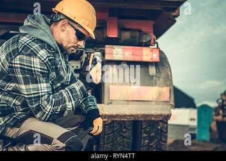 Caucasian Sito in costruzione lavoratore in Hard cappello e occhiali da sole con walkie talkie facendo conversazione con altro edificio membro dell'Equipaggio. Autocarro con cassone ribaltabile in th Foto Stock