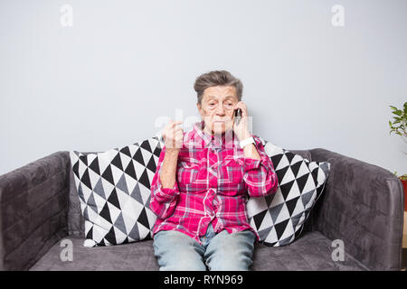 Una vecchia donna anziana nonna con i capelli grigi si siede a casa sul lettino usando il telefono a mano, una conversazione telefonica di sentire le cattive notizie. Emozione Foto Stock