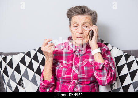 Una vecchia donna anziana nonna con i capelli grigi si siede a casa sul lettino usando il telefono a mano, una conversazione telefonica di sentire le cattive notizie. Emozione Foto Stock