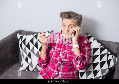 Una vecchia donna anziana nonna con i capelli grigi si siede a casa sul lettino usando il telefono a mano, una conversazione telefonica di sentire le cattive notizie. Emozione Foto Stock
