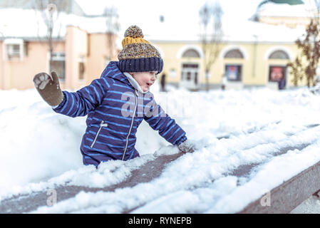 Un bambino di 3-6 anni svolge in inverno nella città, felice divertendosi snowballs, neve raccolta dal banco, sentimenti di gioia e di Foto Stock