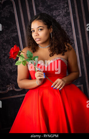 Uno splendido, biracial high school girl senior indossando red party dress il trattenimento di una rosa rossa Foto Stock